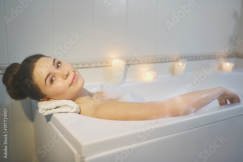 Young girl is resting in a bathtub with foam in bethroom with candles.