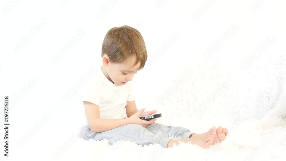 Happy kid playing with a smartphone on a white background. A child is sitting on a white sofa. Happy kid playing with a smartphone on a white background. A child is sitting on a white sofa.
