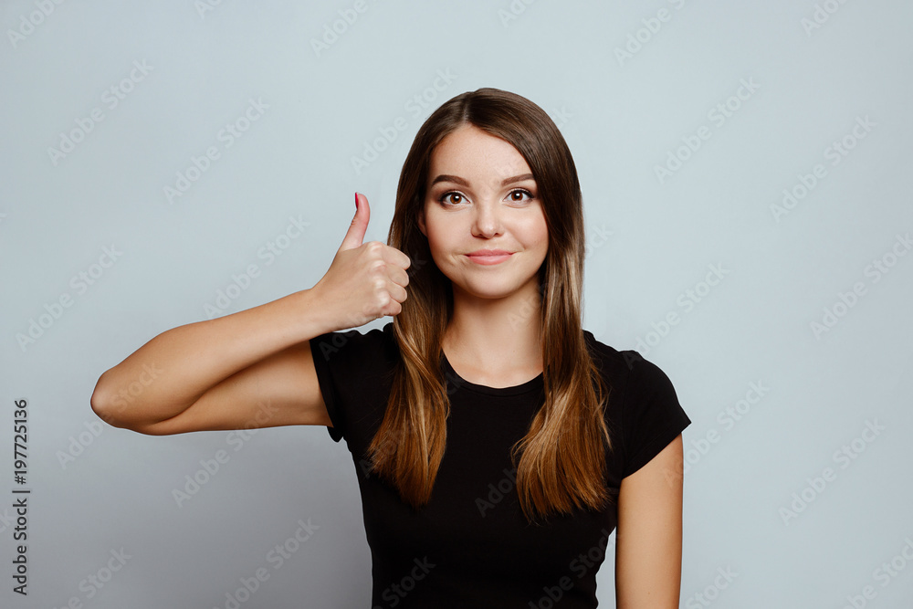 Positive emotions and feelings on white background.Caucasian female with long hair, wearing black T-shirt, showing thumbs up near face and smiling broadly while looking at camera Positive emotions and feelings on white background.Caucasian female with long hair, wearing black T-shirt, showing thumbs up near face and smiling broadly while looking at camera