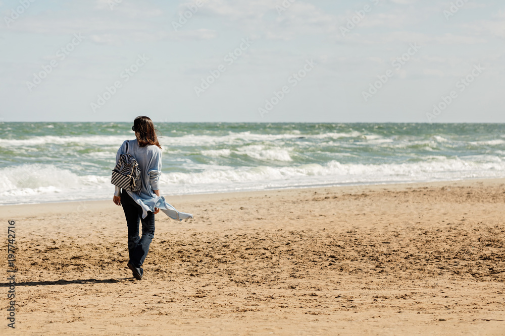 Young woman (brunette) in a light blue cardigan and navy jeans, with a striped backpack, walks along the beach. Young woman (brunette) in a light blue cardigan and navy jeans, with a striped backpack, walks along the beach.