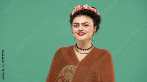 Beautiful portrait of woman with thick eyebrows Frida Kahlo lookalike wearing jewelry and roses in hair smiling with candid artistic, over green background. Stylization concept photo
