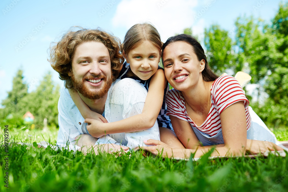 Happy family of parents and daughter looking at camera while relaxing in park on summer day Happy family of parents and daughter looking at camera while relaxing in park on summer day