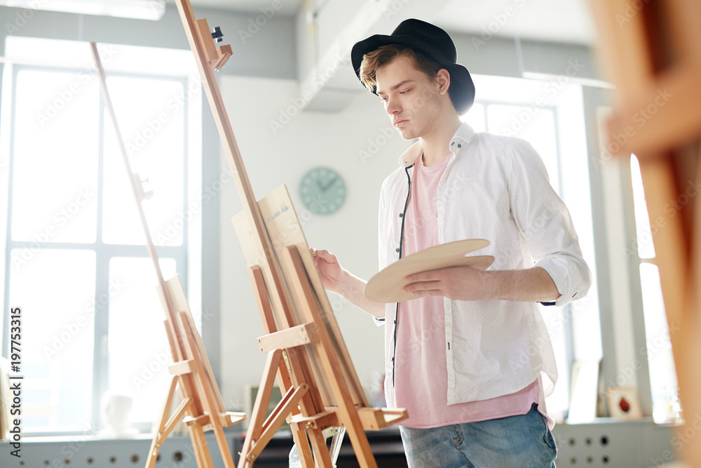 Young guy in casualwear and hat holding palette while standing in front of easel and painting
