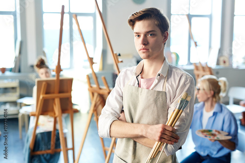 Serious guy in apron holding bunch of paintbrushes at lesson of painting in school of modern arts