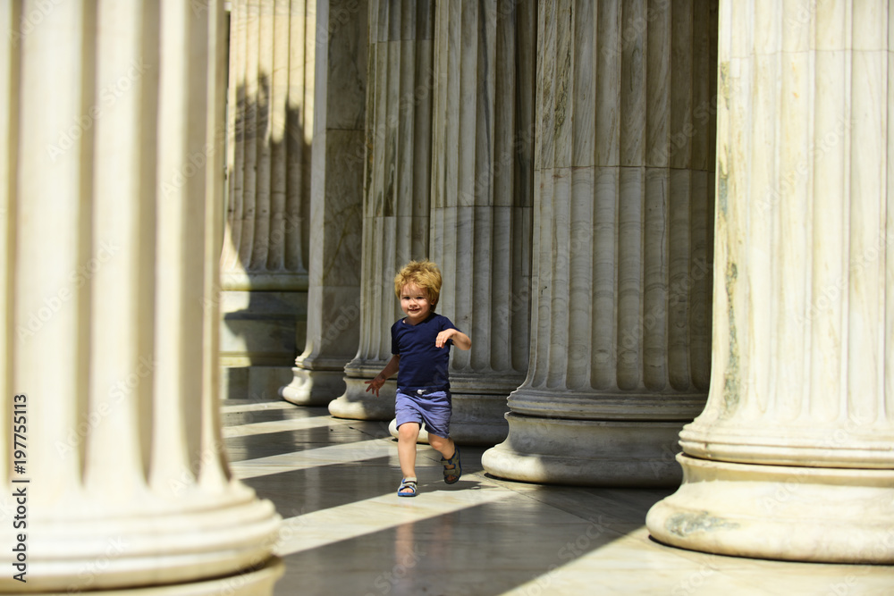Cute child boy running between columns of ancient building in greek style. Kid walks on sunny day, boy on excursion, discovers world around him. Little boy happy to travel, on vacation, holidays.