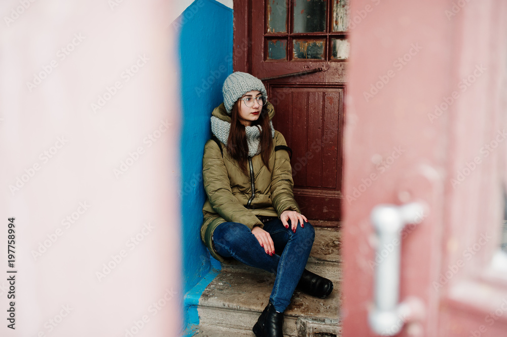 Portrait of brunette girl in gray scarf and hat, glasses against entrance.