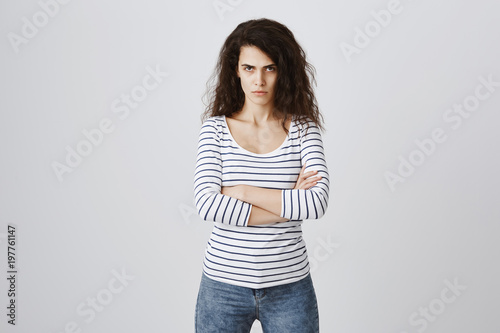 Indoor portrait of angry woman with curly hair, looking from under forehead with frowned eyebrows, crossing hands on chest, being furious and annoyed, showing contempt and dislike with serious face