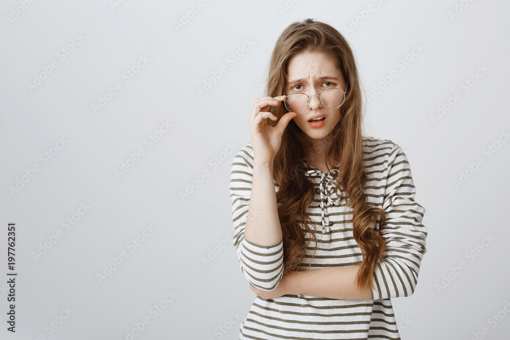 Fototapeta premium Are you kidding me. Studio shot of good-looking puzzled girl with fair hair, frowning, looking from under forehead, taking off glasses and looking with contempt and dislike at camera over gray wall