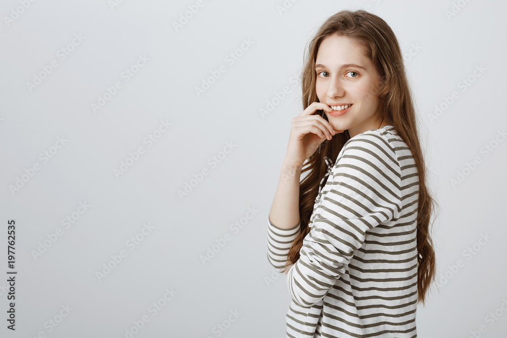Intrigued girl has something curious on mind. Portrait of attractive ordinary female student standing in profile and gazing at camera, holding hand on lip, thinking or wanting something over gray wall Intrigued girl has something curious on mind. Portrait of attractive ordinary female student standing in profile and gazing at camera, holding hand on lip, thinking or wanting something over gray wall
