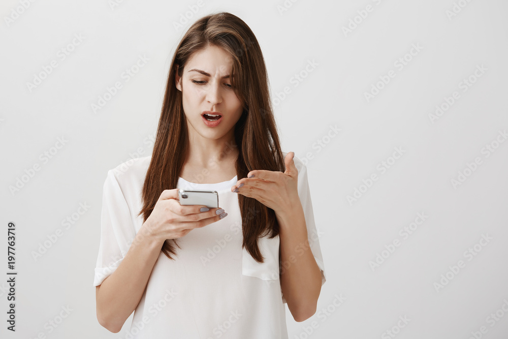 Woman received absurd message. Portrait of displeased annoyed good-looking girl holding smartphone and pointing at screen with palm, complaining, being angry at friend who wrote nasty things Woman received absurd message. Portrait of displeased annoyed good-looking girl holding smartphone and pointing at screen with palm, complaining, being angry at friend who wrote nasty things