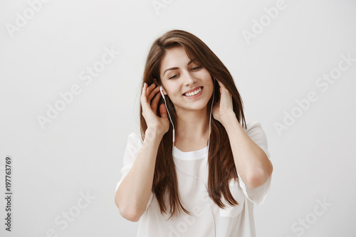 Music has own flavour. Portrait of tender young european female student listening songs in earphones, holding hands on earbuds, smiling broadly, looking down with pleased look, enjoying great sound