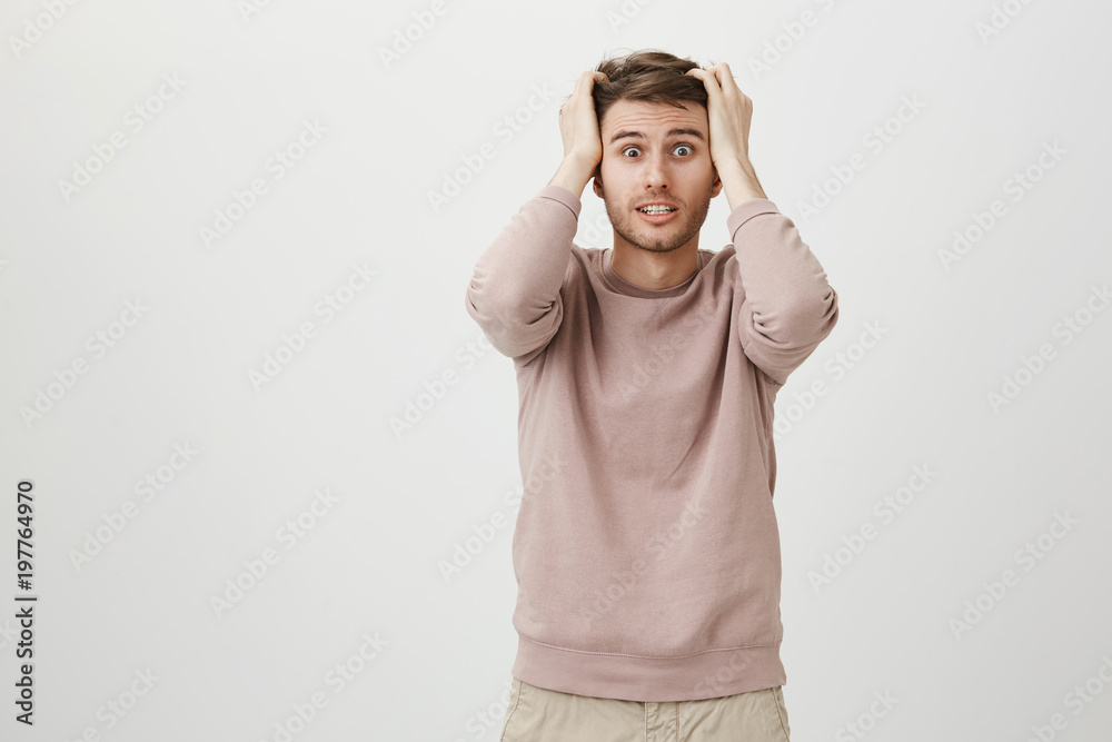 Anxious and worried young good-looking man holding hands on head, looking with popped eyes on something terrifying, standing over gray background. Kitchen caught on fire while he was opening door Anxious and worried young good-looking man holding hands on head, looking with popped eyes on something terrifying, standing over gray background. Kitchen caught on fire while he was opening door