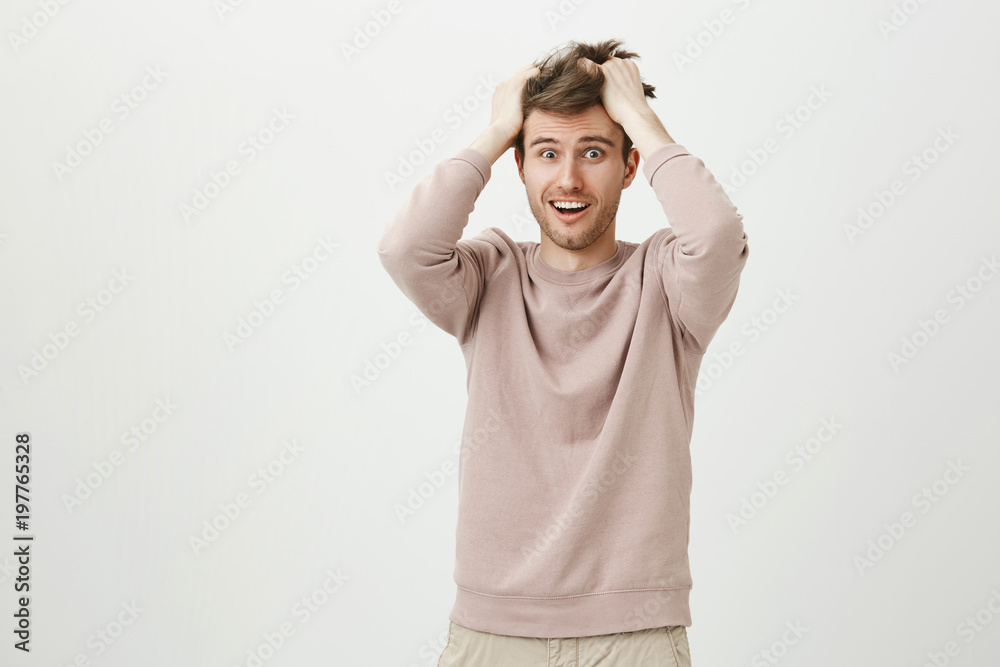 Indoor shot of funny handsome guy messing his hair while rubbing head, expressing devastation and acting crazy, standing over gray background. Guy is shocked after seeing home much work is left Indoor shot of funny handsome guy messing his hair while rubbing head, expressing devastation and acting crazy, standing over gray background. Guy is shocked after seeing home much work is left
