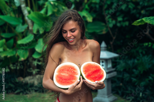 close up portrait of cute smiling brunette girl with a sporty body holds watermelons in her hands and covers her breasts. Photoshoot of a beautiful model with fruits near green palms on background in