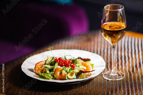 A plate with a beautiful appetizing salad of fresh vegetables  shrimps  quail eggs and lingonberries is on the table with a drink in a wine glass.