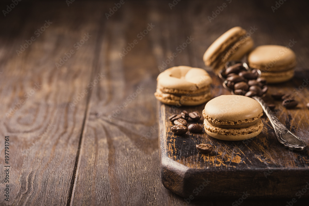 French coffee macarons with ganache filling with coffee beans on old wooden board on vintage background. Holidays food concept with copy space. French coffee macarons with ganache filling with coffee beans on old wooden board on vintage background. Holidays food concept with copy space.