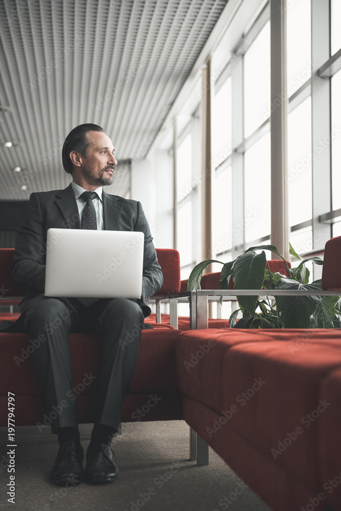 Peaceful mature male in suit sitting on sofa with laptop. Copy space in right side