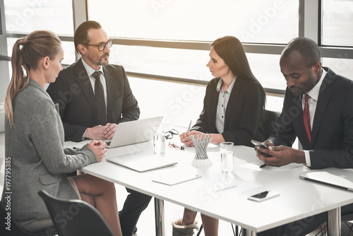 Men and women discussing important questions while taking seat around office desk. Gadgets are on table