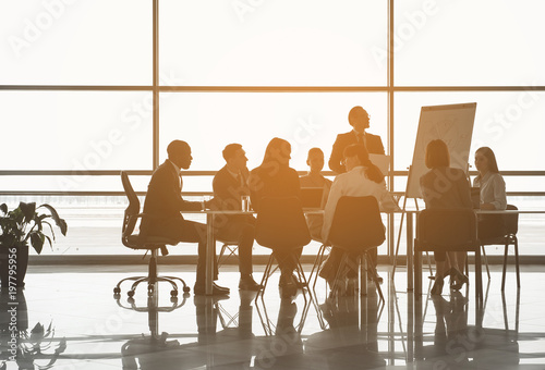Photos Group of people sitting at the office table while their partner standing near board with chart