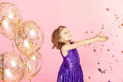 Happy young girl in purple glittering dress celebrating with hands up and smile or skreaming with joy while cathcing confetti, having fun on pink background. photo