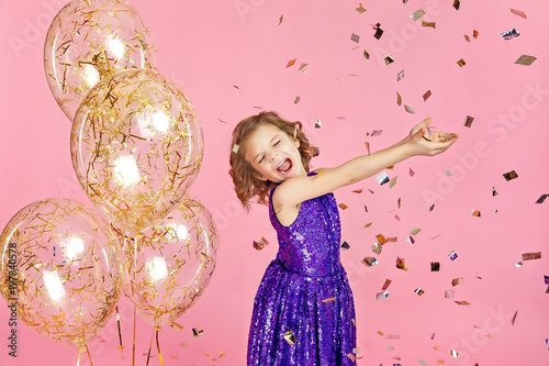 Happy young girl in purple glittering dress celebrating with hands up and smile or skreaming with joy while cathcing confetti, having fun on pink background. photo