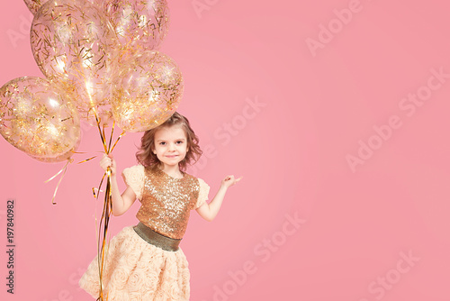 Happy little girl in glittering dress looking at camera on pink background and holding balloons, pointing finger. Copyspace photo