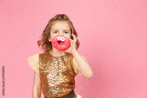 Surprised little girl in glittering dress standing with donuts posing on pink background.