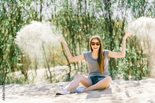 Amazing young girl siiting on beach and looking at camera. Fashion portrait of beautiful female playing with sand beyond sea. Pretty cheerful person smiling face. Traveler relaxing at nature outdoor.