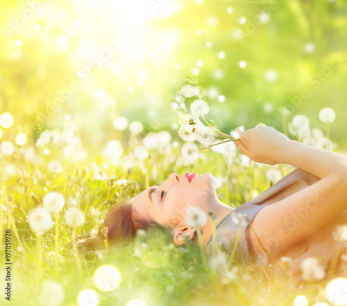 Beautiful young woman lying on the field in green grass and blowing dandelion. Healthy smiling girl on spring lawn. Allergy free concept