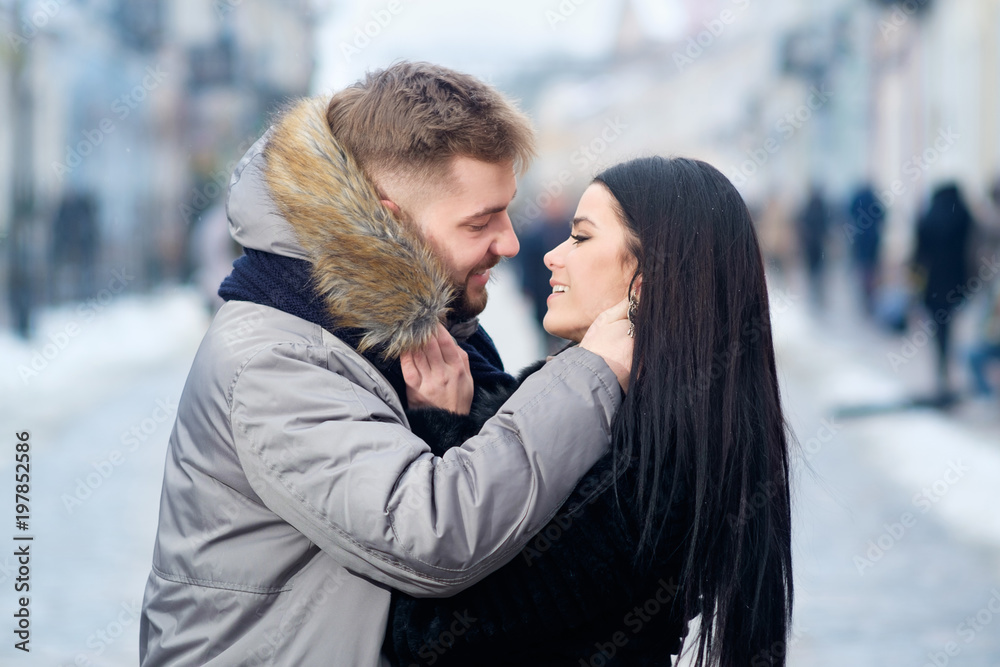 Fototapeta premium young happy lovely caucasian couple hug and kiss each other while walking in the street of town in winter under snowfall