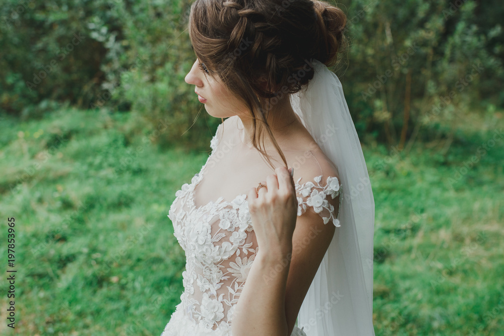 Beautiful bride with elegant simple hairdo is standing in the forest. Low open back white dress with flowers and tulle veil. Stylish hairstyle in outdoors romantic wedding photo. Rustic details. Beautiful bride with elegant simple hairdo is standing in the forest. Low open back white dress with flowers and tulle veil. Stylish hairstyle in outdoors romantic wedding photo. Rustic details.