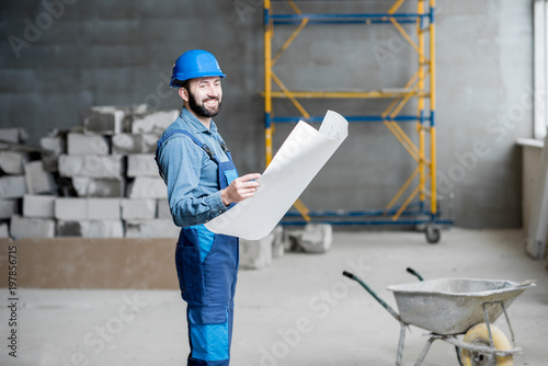 Builder or foreman in working uniform expertising the structure standing with blueprint at the construction site indoors