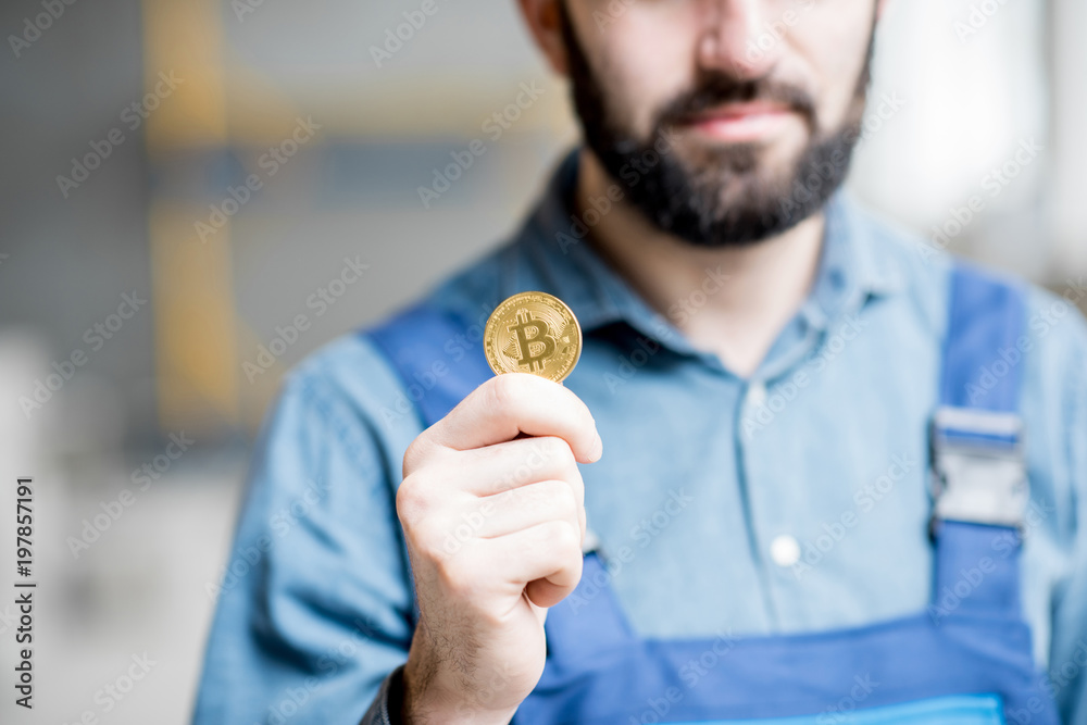 Builder in working uniform holding cryptocoin as a salary standing at the construction site indoors Builder in working uniform holding cryptocoin as a salary standing at the construction site indoors