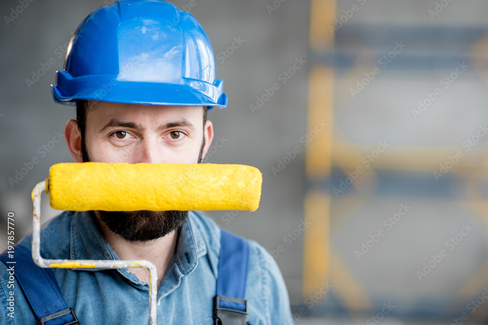 Portrait of a builder in working uniform holding yellow paint roll and grinding tool indoors Portrait of a builder in working uniform holding yellow paint roll and grinding tool indoors