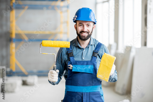 Portrait of a builder in working uniform holding yellow paint roll and grinding tool indoors photo
