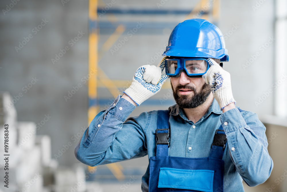 Close-up portrait of a handsome bearded builder with protective glasses and helmet indoors - obrazy, fototapety, plakaty 