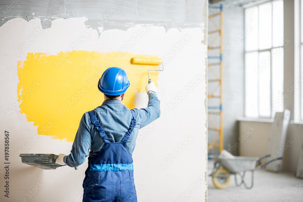 Workman in uniform painting wall with yellow paint at the construction site indoors - obrazy, fototapety, plakaty 