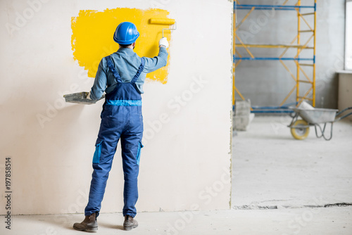 Fototapeta Naklejka Na Ścianę i Meble -  Workman in uniform painting wall with yellow paint at the construction site indoors