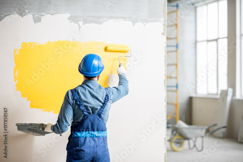 Workman in uniform painting wall with yellow paint at the construction site indoors photo