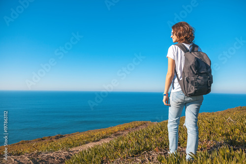 Young attractive girl in a white T-shirt and jeans walking on a background of blue sea and sky, summer travel and adventure, vacation concept