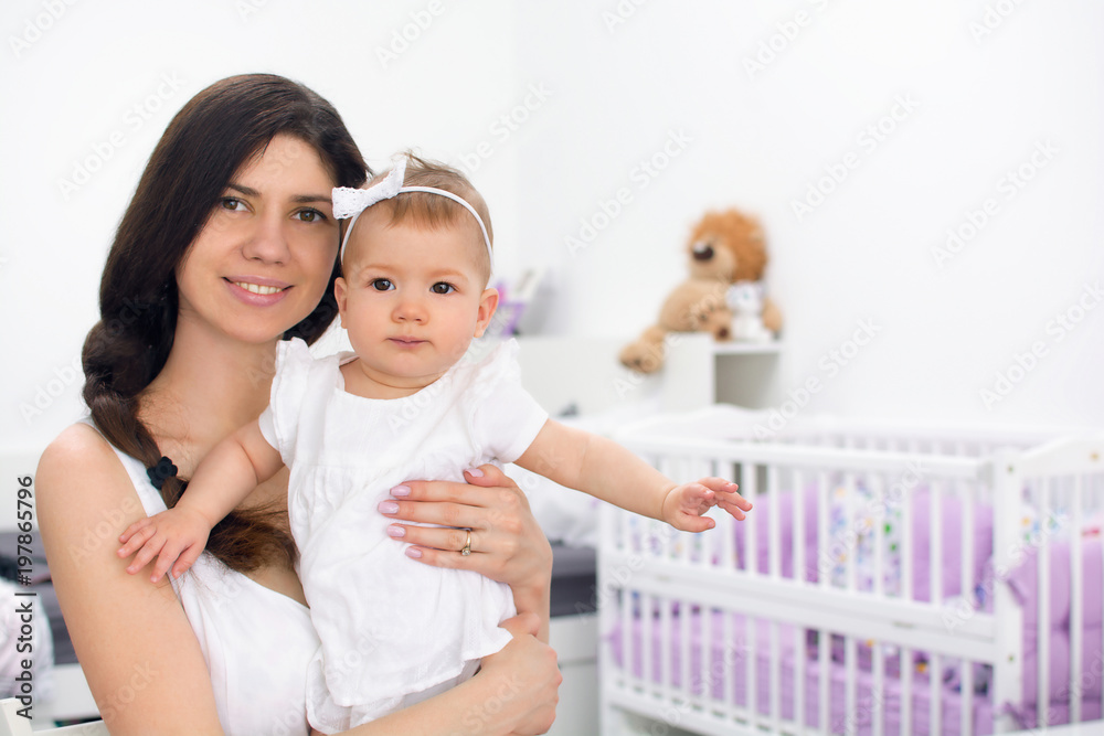 Young mother smiling and holding her daughter child. Woman and baby girl feeling happy in a white bedroom with toys and crib. Nursery interior. Family at home
