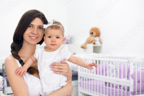 Obraz na płótnie Young mother smiling and holding her daughter child. Woman and baby girl feeling happy in a white bedroom with toys and crib. Nursery interior. Family at home w salonie