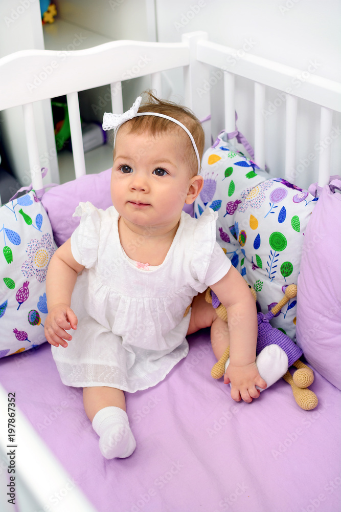 Portrait of 7 month old baby girl sitting in white crib in light room