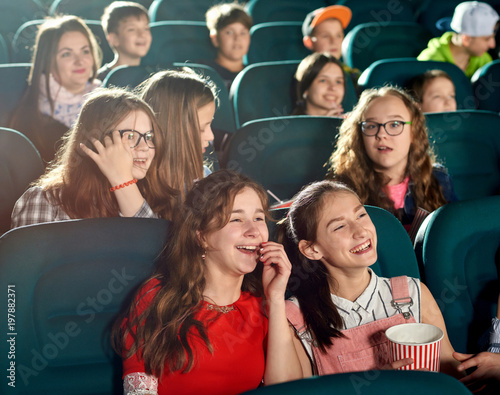 Happy girls laughing by watching interesting movie in the cinema. They smiling, looking satisfied. There are many other emotional children on the background.