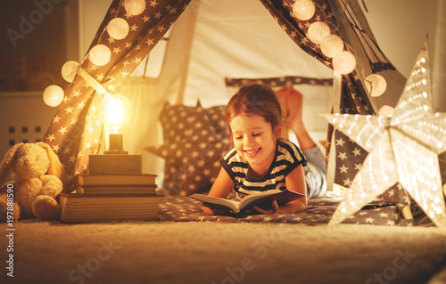 happy child girl laughing and reading book in dark in tent at home