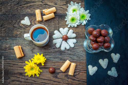 Fototapeta Naklejka Na Ścianę i Meble -  Sweet breakfast or snack. White cup with freshly brewed coffee, chocolate candies, heart shape cookies, waffle rolls, white and yellow flowers on a wooden table.