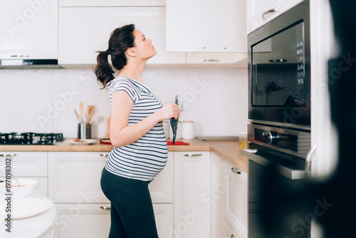 Young pregnant woman cooking in her kitchen standing and looking in cabinets near stove
