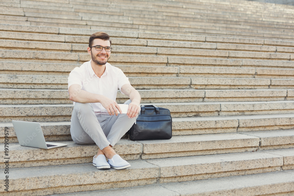 Cheerful young businessman using phone and laptop on stairs