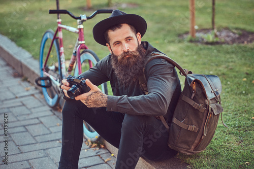 A handsome hipster traveler with a stylish beard and tattoo on his arms dressed in casual clothes and hat with a bag, using the smartphone, sitting on the sidewalk, resting after a bike ride.