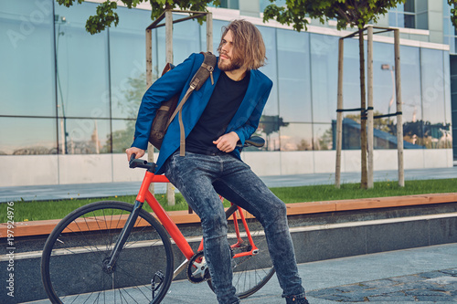 A handsome bearded man in stylish jeans and jacket with a backpack sitting on a bicycle in the city.